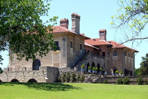 A large stone mansion with a red tile roof, surrounded by greenery and featuring multiple chimneys and arched windows.