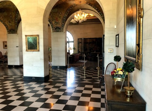 Elegant interior with arched doorways, checkered floor, and decorative ceiling, featuring artwork and potted plants.