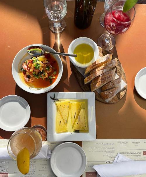 A table set with a colorful seafood dish, olive oil, bread, cheese, and drinks in glasses.
