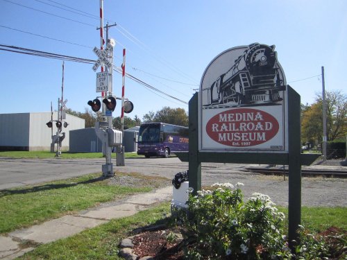 Sign for Medina Railroad Museum with a train crossing signal and a bus in the background on a clear day.
