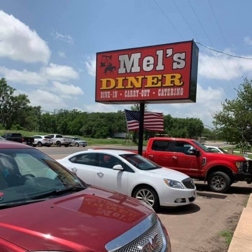 Sign for Mel's Diner with "DINE-IN, CARRY-OUT, CATERING" and parked cars in front, including a red truck and a white car.