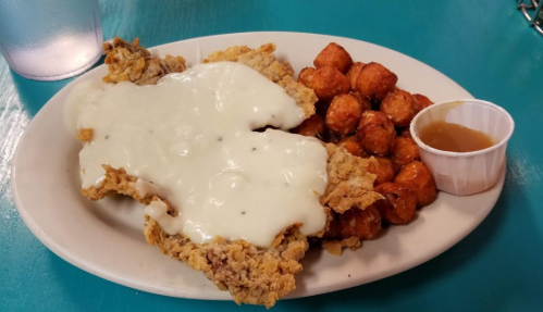 A plate of fried chicken smothered in white gravy, served with tater tots and a small cup of dipping sauce.