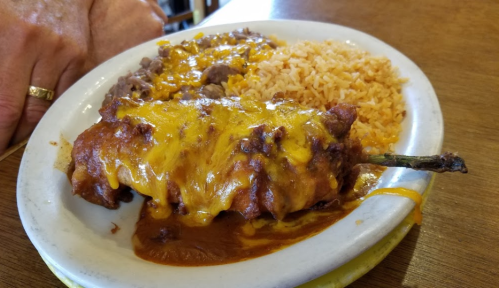 A plate of food featuring a cheese-covered enchilada, rice, and refried beans on a wooden table.