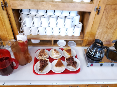 A tray with slices of pie on plates, coffee pot, and pitchers, next to a shelf of stacked mugs and cups.