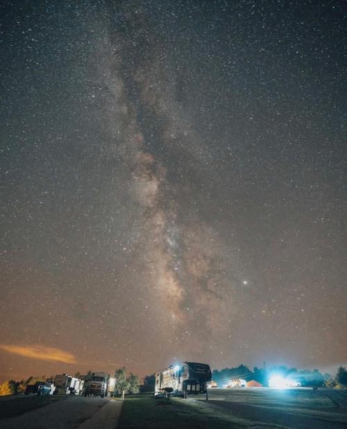 A starry night sky with the Milky Way visible above a campsite, featuring RVs and trees in the foreground.
