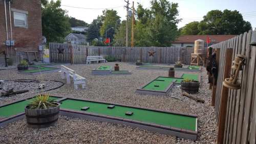 A mini-golf course with green turf, surrounded by gravel, wooden fences, and rustic decor in a sunny outdoor setting.