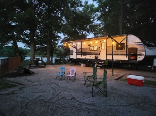 A camper trailer parked by a lake, surrounded by chairs and a cooler, with string lights glowing in the evening.
