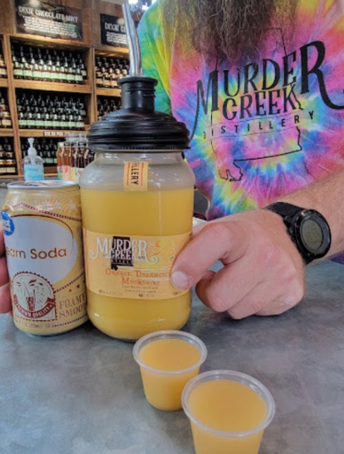 A person in a colorful shirt holds a drink and a can of cream soda, with two small cups of orange drink on the table.