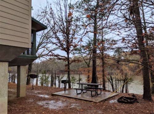 A wooden deck with a picnic table near a lake, surrounded by trees and a house on stilts. Overcast sky.