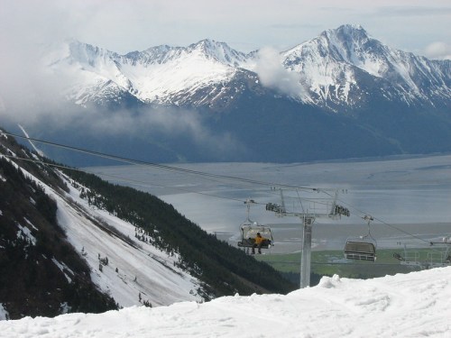 A ski lift ascends a snowy mountain, with majestic peaks and a calm body of water in the background.