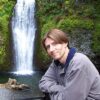 A person smiles by a serene waterfall surrounded by lush greenery.
