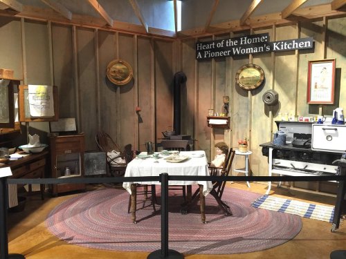 A pioneer woman's kitchen display featuring a table set with dishes, a stove, and rustic decor in a wooden room.