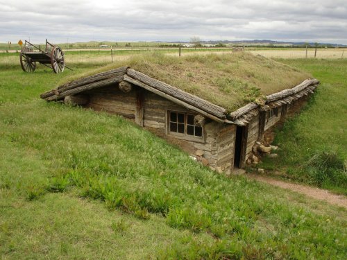 A rustic, grass-roofed cabin made of logs, set in a grassy field with a wooden cart nearby under a cloudy sky.