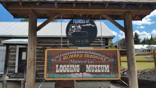 Sign for the J. Howard Bradbury Memorial Logging Museum, featuring a wooden structure and a blue sky backdrop.