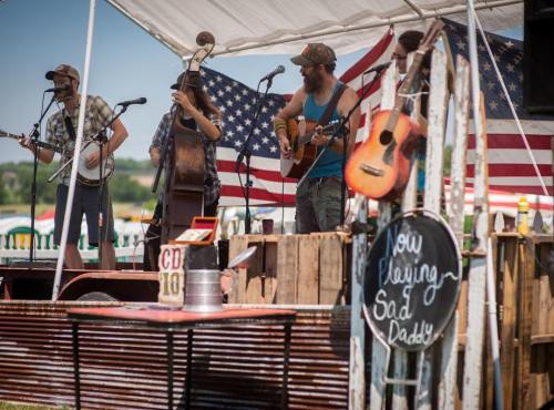 A band performs on stage with an American flag backdrop, featuring guitars and a sign that reads "Now Playing: Sad Daddy."