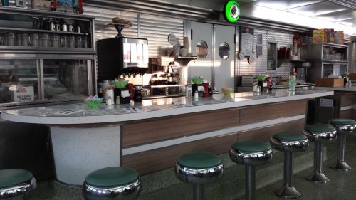 A retro diner interior featuring a long counter with green stools, coffee machines, and vintage decor.
