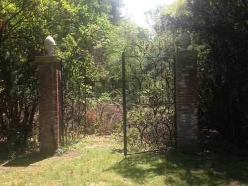 Wrought iron gates open to a lush, green garden surrounded by trees and shrubs. Sunlight filters through the foliage.