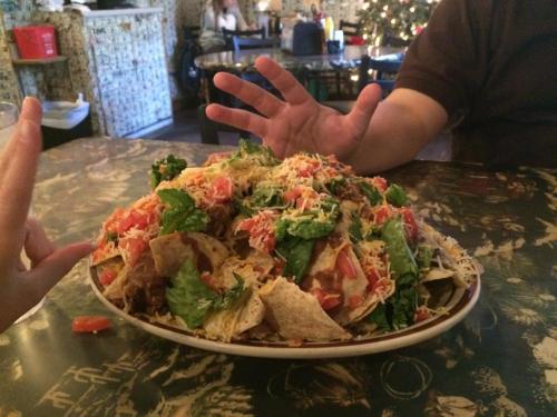 A large plate of nachos topped with lettuce, tomatoes, and cheese, with hands reaching towards it on a patterned table.
