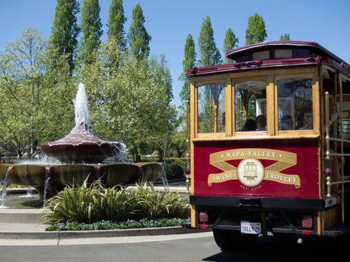 A red trolley with "Napa Valley Wine Trolley" signage near a fountain and lush greenery on a sunny day.