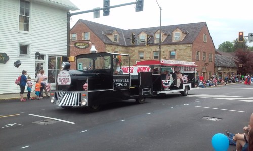 A parade scene featuring a black train-like float and a red vehicle, with spectators lining the street.