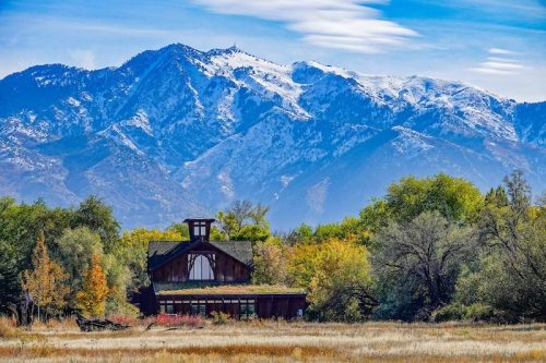 A rustic wooden building surrounded by colorful trees, with snow-capped mountains in the background under a clear blue sky.