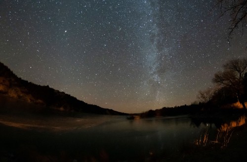 A serene night sky filled with stars and the Milky Way over a calm river surrounded by hills and trees.