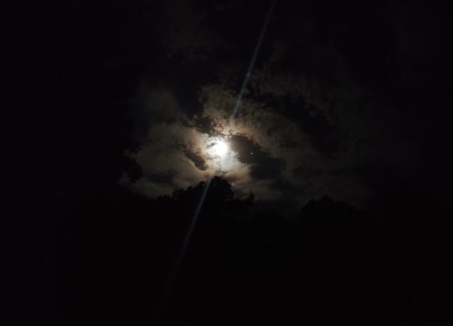 A bright moon partially obscured by clouds, casting a soft glow in a dark night sky.