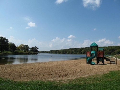 A sandy beach by a calm lake, featuring a colorful playground structure under a blue sky with fluffy clouds.