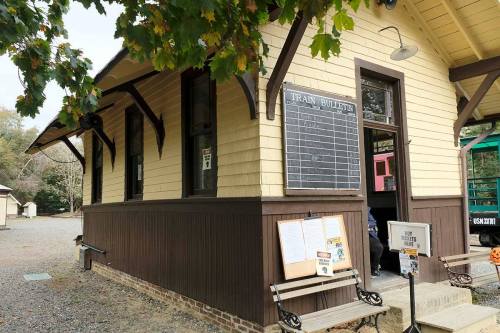 Historic train station with a bulletin board, surrounded by trees and a gravel path.