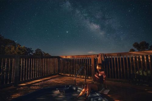 A person sits by a hot tub on a deck, gazing at a starry sky filled with the Milky Way.