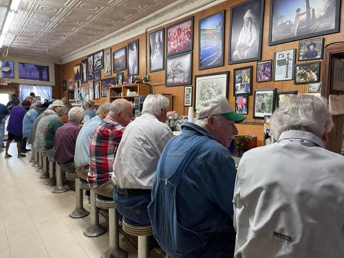 A row of elderly men sitting at a diner counter, surrounded by framed photos on the wall.