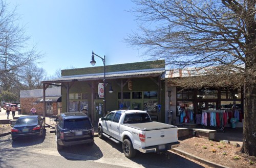 A street view of a small shopping area with parked cars and storefronts under a clear blue sky.