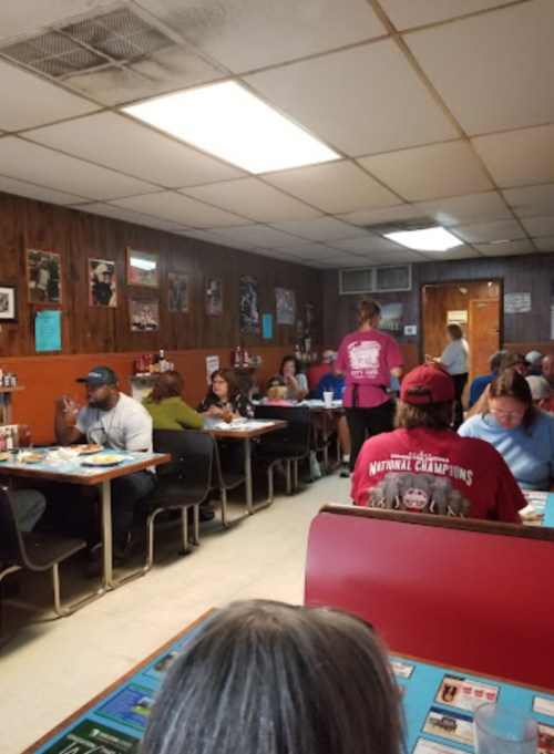 A busy diner with patrons seated at tables, a server in a pink shirt, and wooden paneling on the walls.