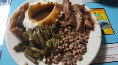 A plate of food featuring mashed potatoes with gravy, pulled pork, green beans, black-eyed peas, and a fried item.