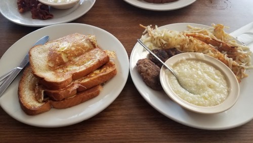 A plate with French toast, a side of sausage, hash browns, and a bowl of grits on a wooden table.