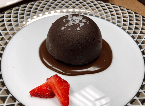 A round chocolate dessert topped with sea salt, served on a chocolate sauce with fresh strawberry slices.