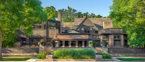 A large, modern house surrounded by lush greenery and trees, featuring a mix of architectural styles and textures.