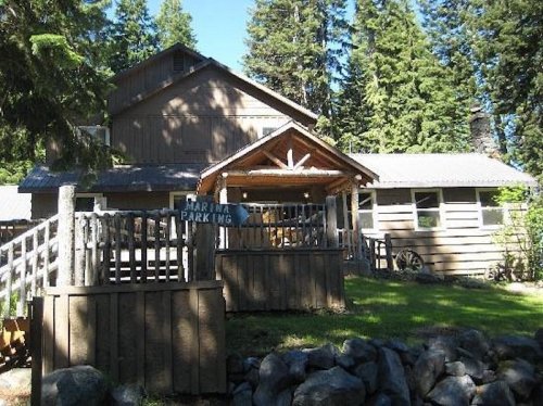A rustic wooden building surrounded by tall trees, featuring a sign for parking and a stone wall in the foreground.