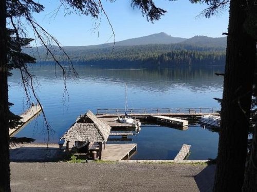 A serene lake view with a wooden dock, surrounded by trees and mountains under a clear blue sky.