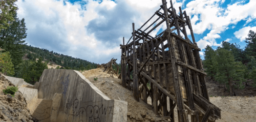 Abandoned wooden structure on a hillside, surrounded by trees and rocky terrain under a cloudy sky.