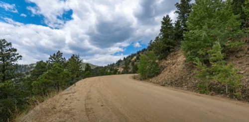 A winding dirt road surrounded by green trees and hills under a cloudy sky.