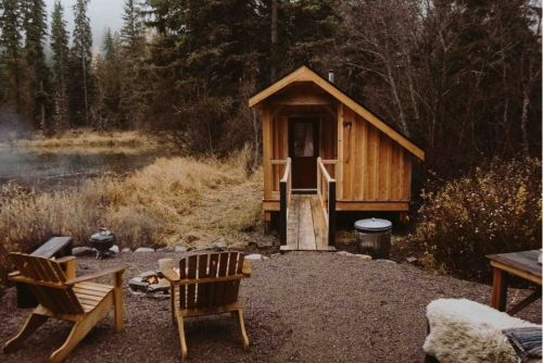 A cozy wooden cabin surrounded by trees, with a fire pit and chairs in the foreground, set in a tranquil outdoor setting.