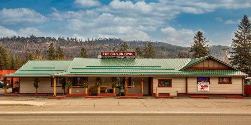 A rustic building with a green roof, "The Silver Spur" sign, surrounded by trees and mountains under a cloudy sky.
