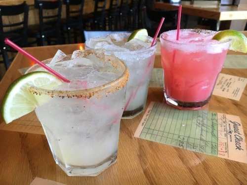 Three colorful cocktails with lime wedges and straws on a wooden table, with menus in the background.