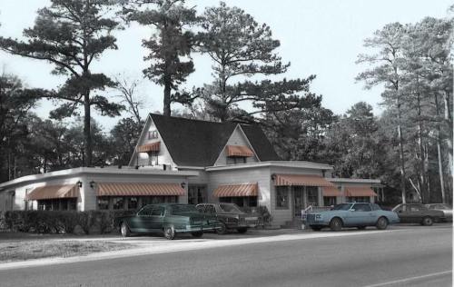 A vintage diner with orange awnings, surrounded by tall trees and parked classic cars on a quiet street.