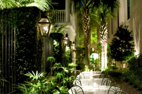 A beautifully lit outdoor dining area with tables surrounded by lush greenery and palm trees.