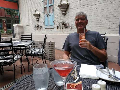A man sits at an outdoor table, enjoying a cocktail, with a martini and water glass in front of him.