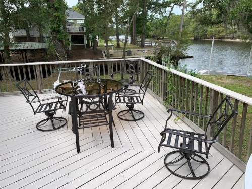A wooden deck with a round table and six metal chairs, overlooking a calm river surrounded by trees.