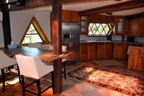 A cozy kitchen with wooden cabinets, a dining table, and a patterned rug, featuring large windows and modern appliances.
