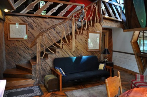 Cozy interior of a wooden house featuring a staircase, a sofa, and rustic decor. Natural light filters in through windows.